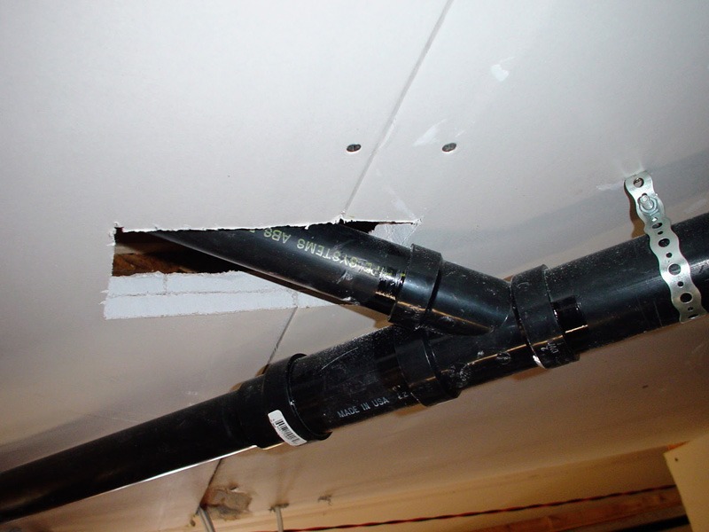 Black ABS drain pipes running below a white drywall ceiling. One pipe passes up through a ragged, oversized cut in the drywall with no firestopping installed.