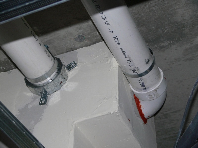 Two white 4-inch PVC pipes beneath a concrete ceiling. One has a metal firestop collar fitted at a rough, white-patched enclosure around the penetration. The other pipe runs into a boxed section, with red sealant visible near its elbow.
