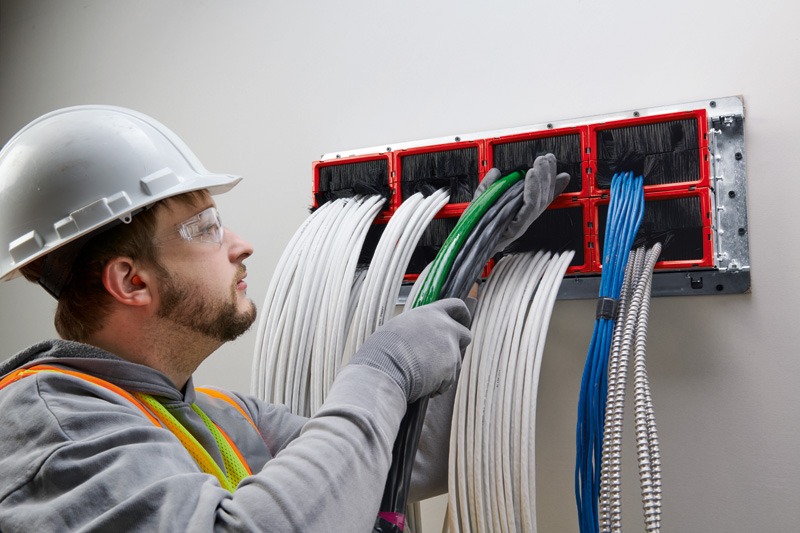 Worker in a white hard hat, safety glasses, and high-visibility vest feeding bundles of white, green, blue, and metal-clad cables through a wall-mounted modular cable pathway device. The device has red frames with black brush-style inserts.