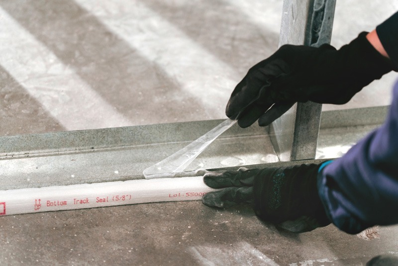 Gloved hands peeling the backing from a white pre-formed firestop strip, labeled “Bottom Track Seal,” along the bottom track of a metal-stud wall on a concrete floor.