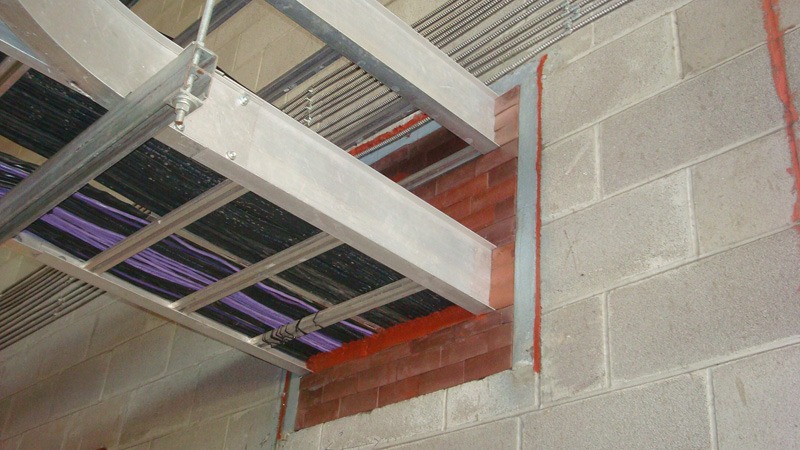 Cable tray carrying black and purple cables through an opening in a concrete block wall. The opening is filled with red-orange firestop material, with a red sealant bead along the edges.
