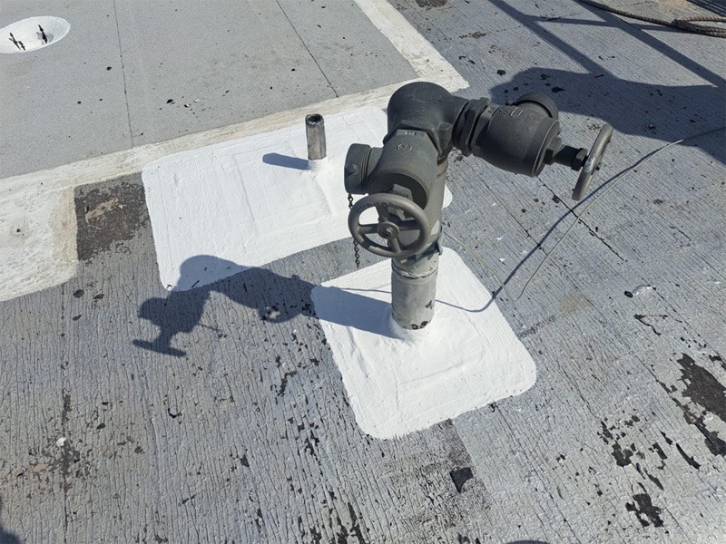 Gray roof surface with a metal pipe and a valve assembly rising from it. White sealant or coating is applied in rough rectangular patches around the base of the valve and a nearby capped pipe, forming flashing that seals the penetrations.