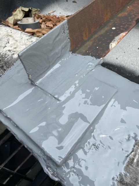 Close-up of a roof curb corner with a wet, glossy gray coating applied over the metal flashing and the adjoining surface. Rust shows on the metal behind the curb, and dry leaves and a roll of tape sit in the background.