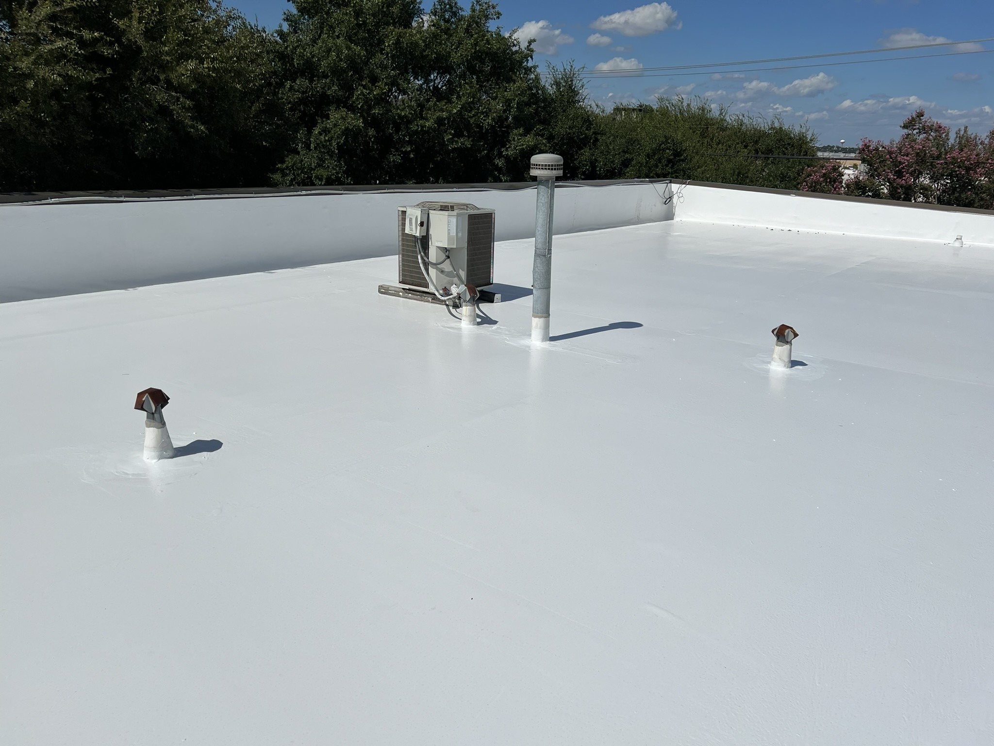 Flat white-coated commercial roof under a blue sky, with a low white parapet wall behind. An HVAC condenser unit, a tall gray vent pipe with a cap, and two rusty-capped vent pipes sit on the roof, each base sealed with white coating, with trees in the background.