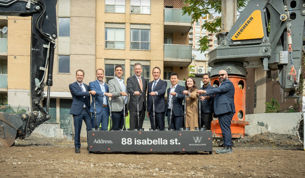 Nine people pose with shovels at a construction site beside heavy machinery and a sign reading “88 isabella st.”