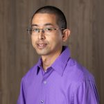 Portrait of a smiling man wearing rectangular glasses and a bright purple collared shirt in front of a wood-paneled background.