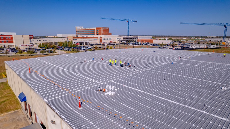 Aerial view of a large commercial building with a gray metal roof, where workers in yellow safety vests apply sealant along the roof seams. Buckets and supplies sit on the roof, and orange safety markers line one edge, with a hospital complex and construction cranes in the background.