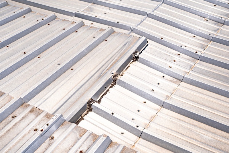 Close-up of a metal roof where two panel sections meet at an angle. The ribbed panels have visible fasteners, and a wide gap along the joint shows deteriorated sealant and debris.
