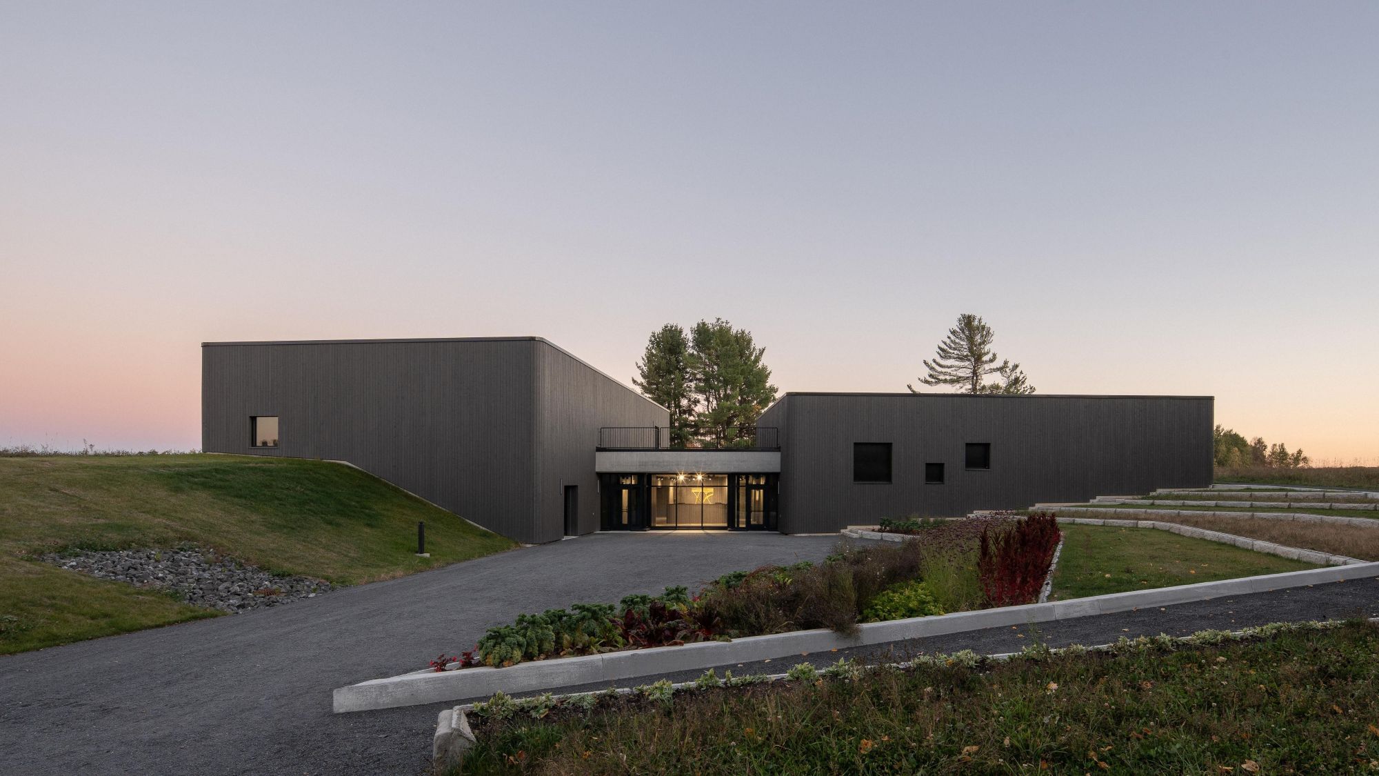 Modern dark-gray building with a recessed, warmly lit glass entrance, surrounded by grassy landscaping and trees at dusk.