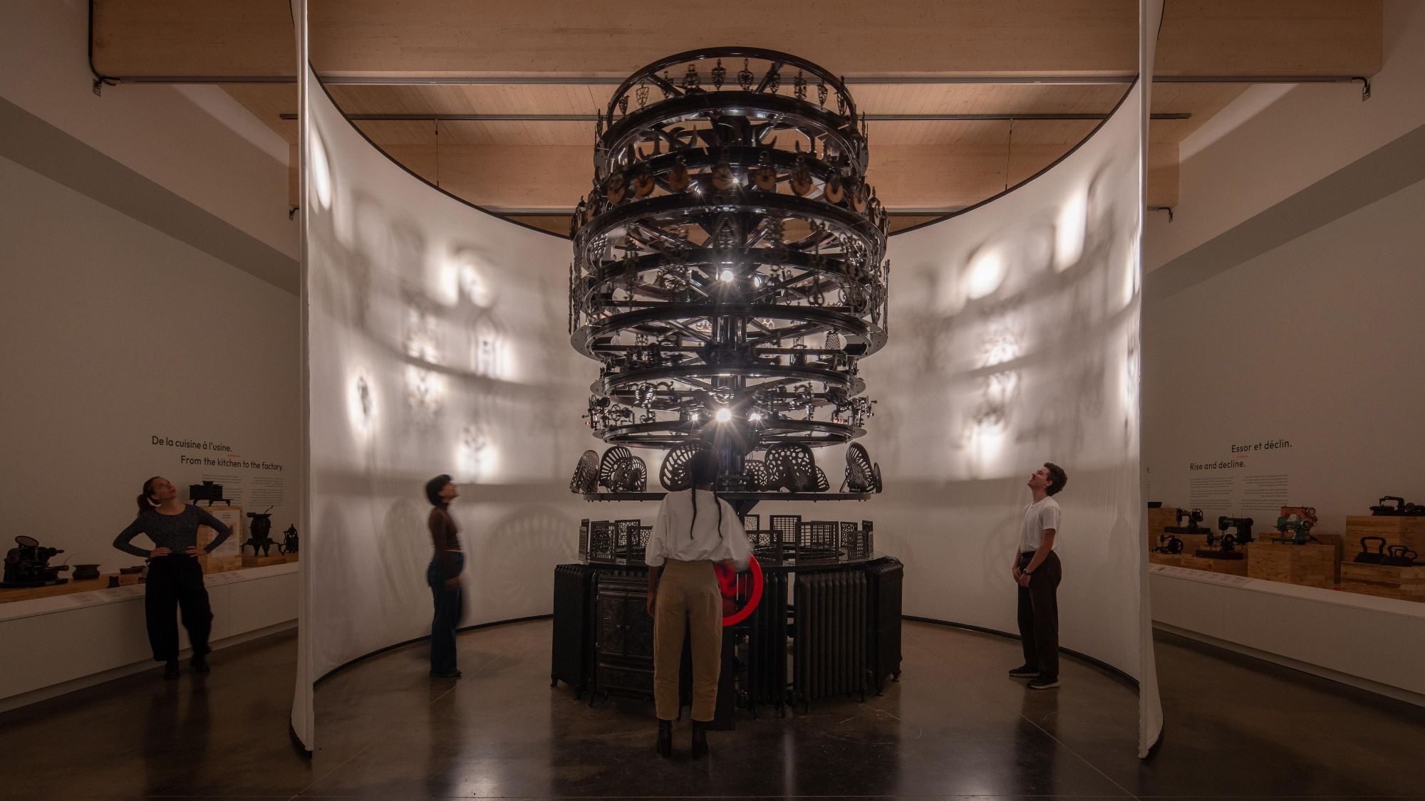 Museum gallery with visitors viewing a large suspended, multi-tiered mechanical sculpture surrounded by historical industrial machines.