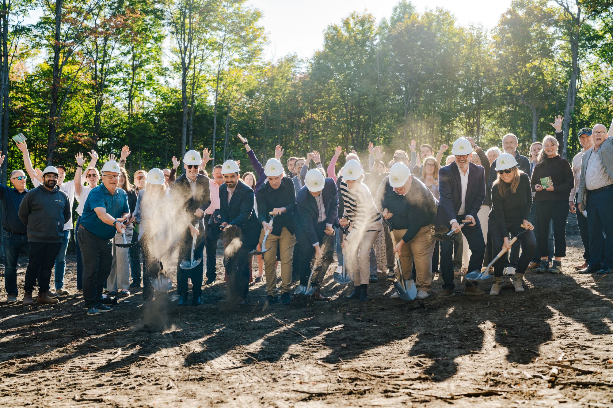 Group wearing hard hats participates in a ceremonial groundbreaking among trees, raising shovels and tossing soil.