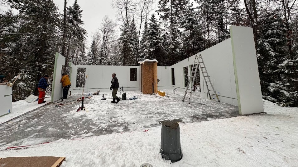 Partially constructed white building in a snowy forest, with workers, a ladder, and construction equipment outside.