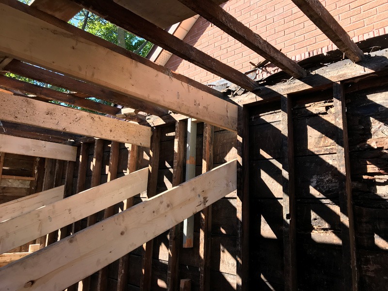 Exposed wooden framing inside a partially dismantled structure, with diagonal braces, missing roof sections, and a red brick wall in the background.