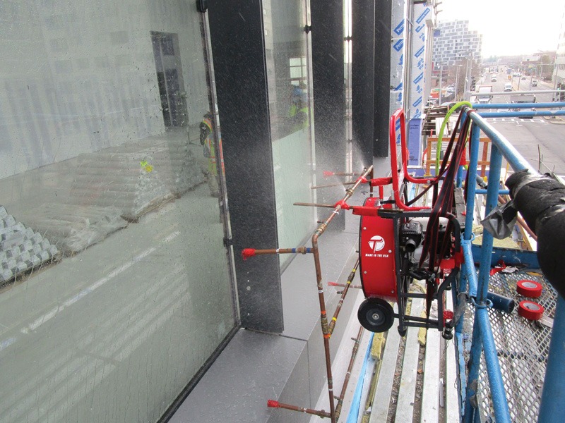 Window-cleaning equipment and hoses on a suspended platform beside a reflective glass building, with a city street visible in the background.