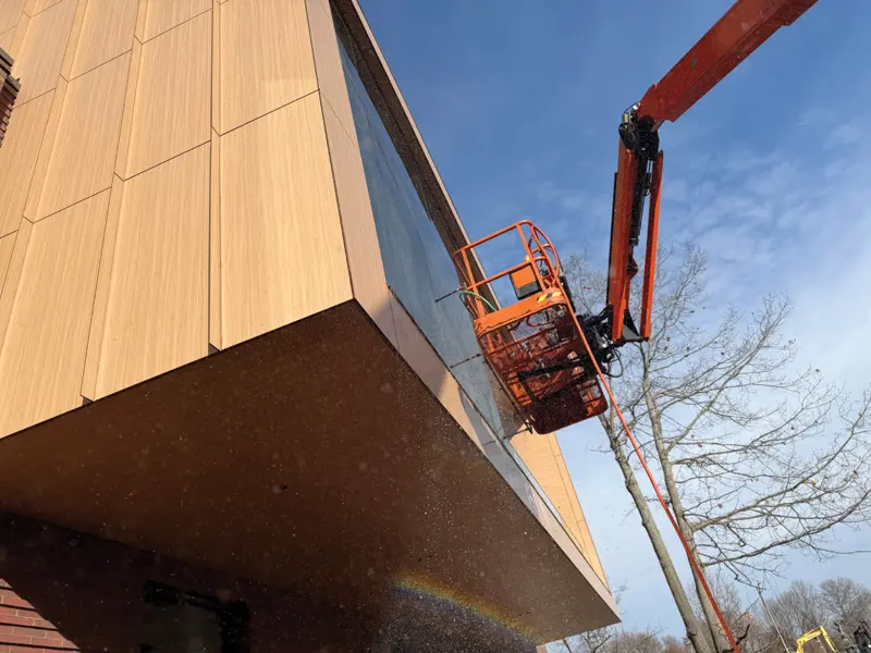 Orange boom lift raised beside the wood-paneled and glass facade of a modern building, viewed from below.