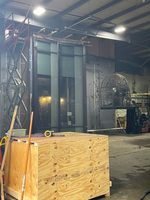 Industrial warehouse with a plywood crate in the foreground, equipment, a scissor lift, and a forklift in the background.