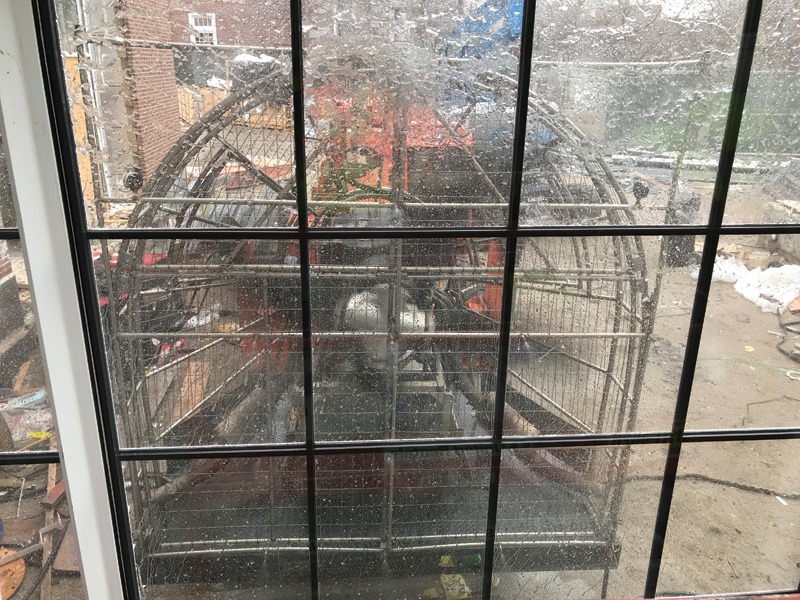 View through a wet, gridded window of metal construction equipment outside, with urban buildings and patches of snow in the background.