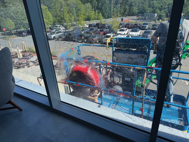 View through a window of outdoor machinery with blue railings, a red circular component, and a parking lot surrounded by trees.