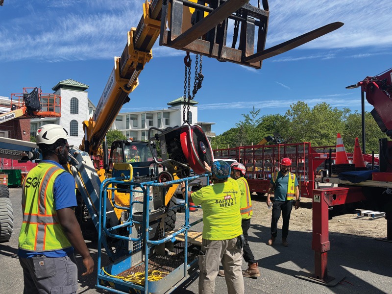 Construction workers in safety gear demonstrate lifting equipment at an outdoor worksite.