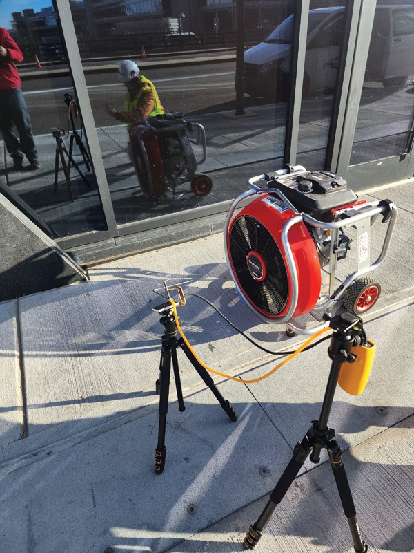 A red portable industrial fan and connected sensor equipment on tripods outside a glass building, with a worker and vehicles reflected in the windows.