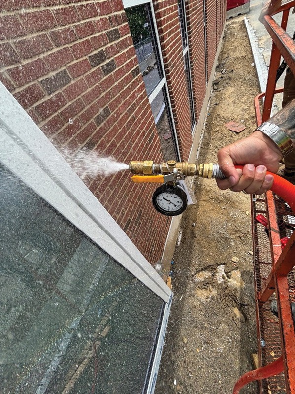 Worker sprays water onto a glass window beside a brick building using a hose with a pressure gauge.