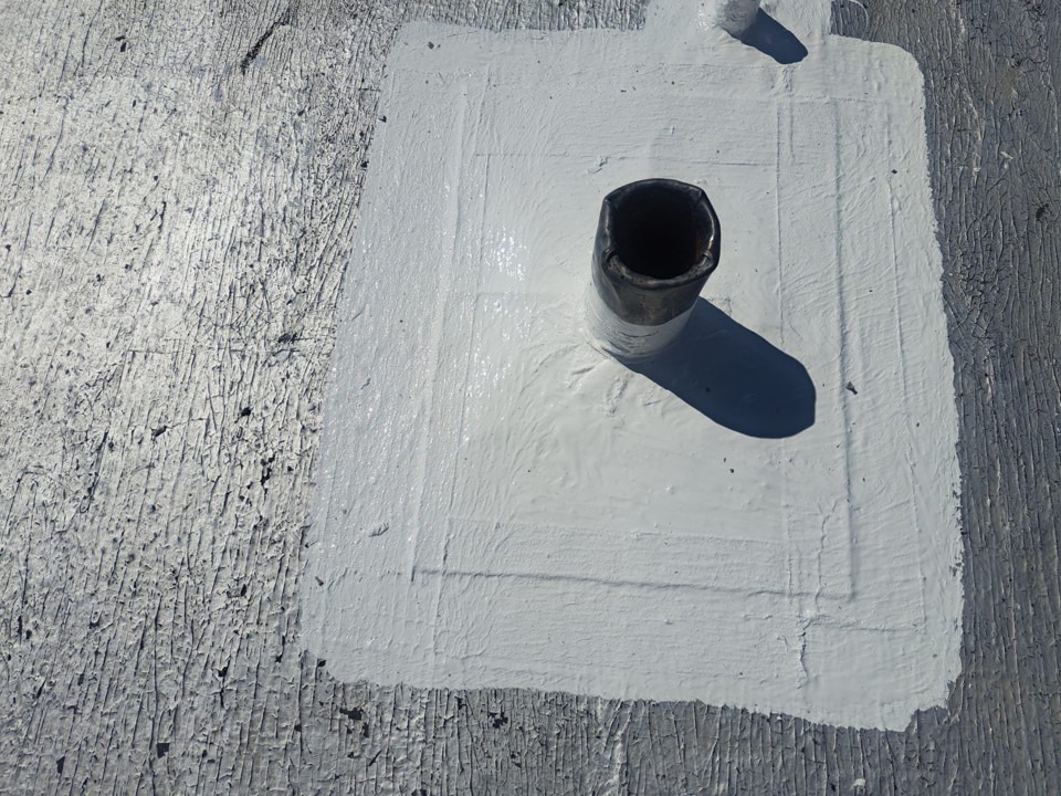 Close-up of a dark pipe penetration rising from a gray roof surface. A square patch of light gray coating surrounds the base of the pipe, with visible brush or trowel marks in the material.