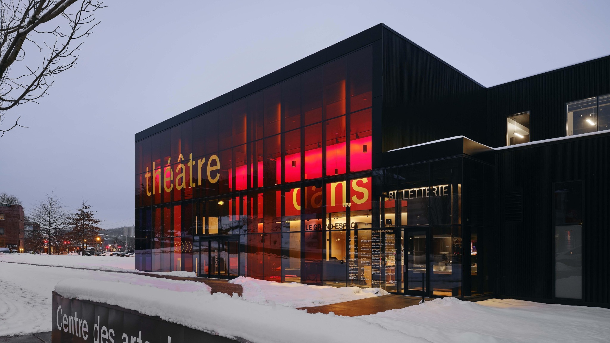 Snow-covered modern arts center with a red-lit glass façade, “BILLETTERIE” entrance, and “Centre des arts” sign.