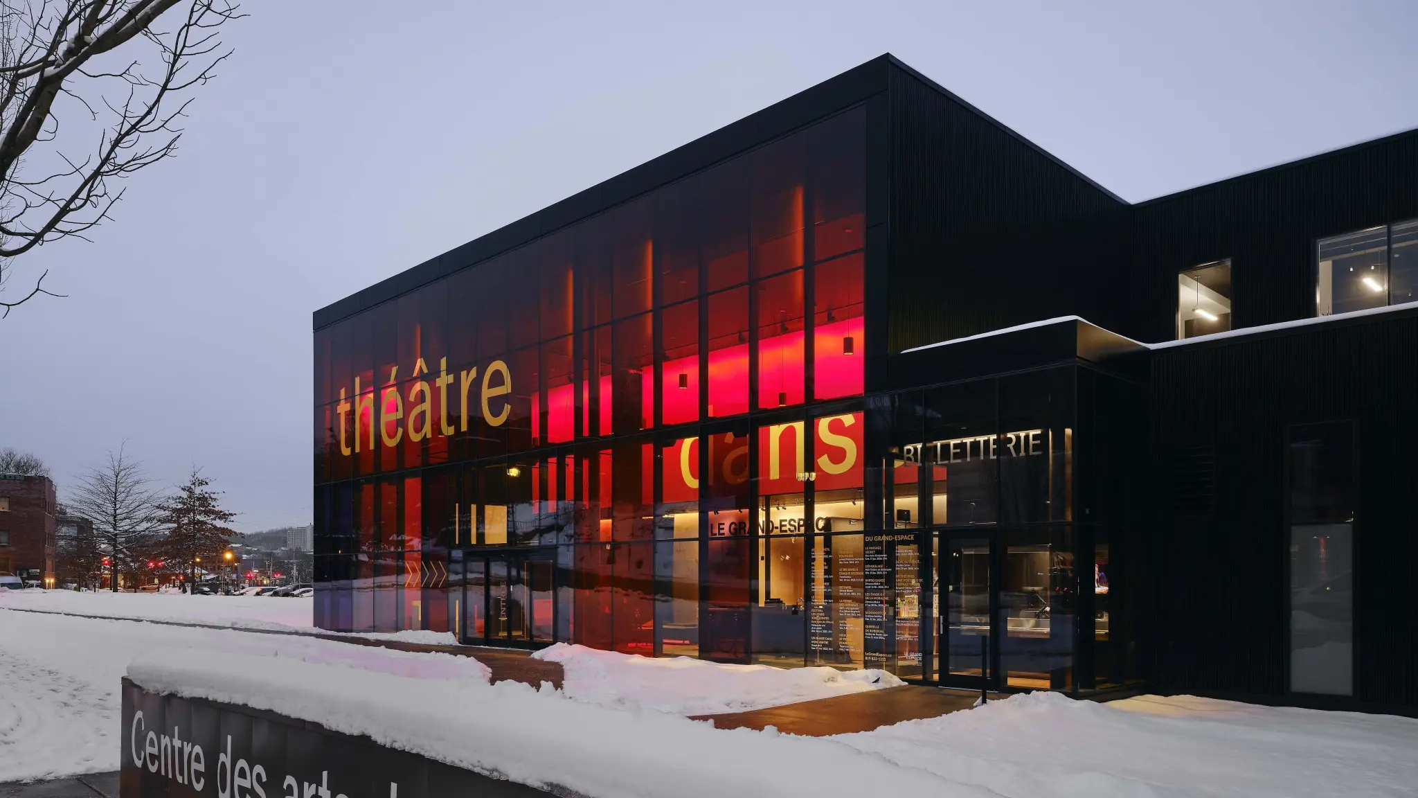 Snow-covered modern arts center with a red-lit glass façade, “BILLETTERIE” entrance, and “Centre des arts” sign.