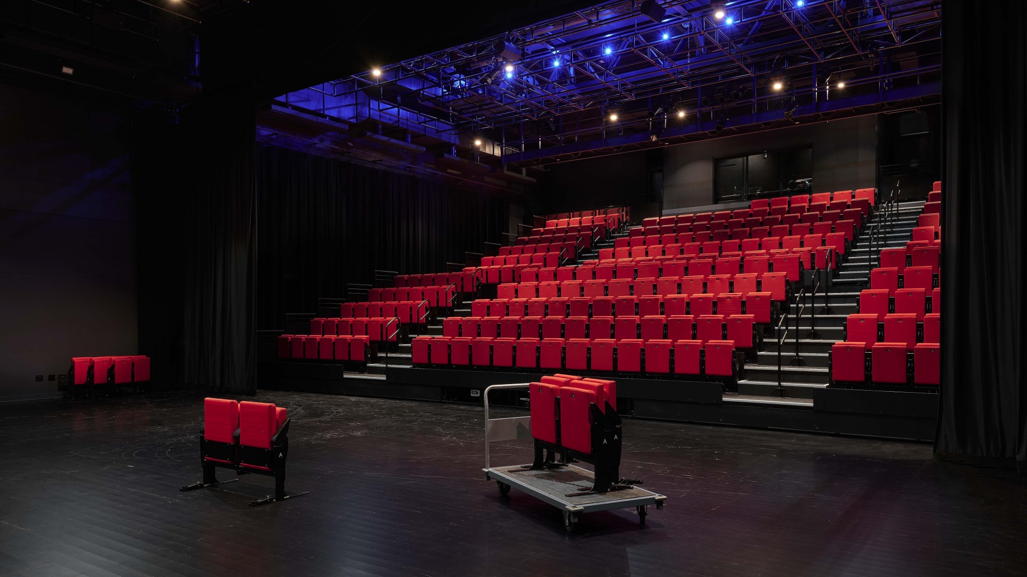 Empty theater auditorium viewed from the stage, with tiered rows of red seats, black curtains, and blue stage lights overhead.