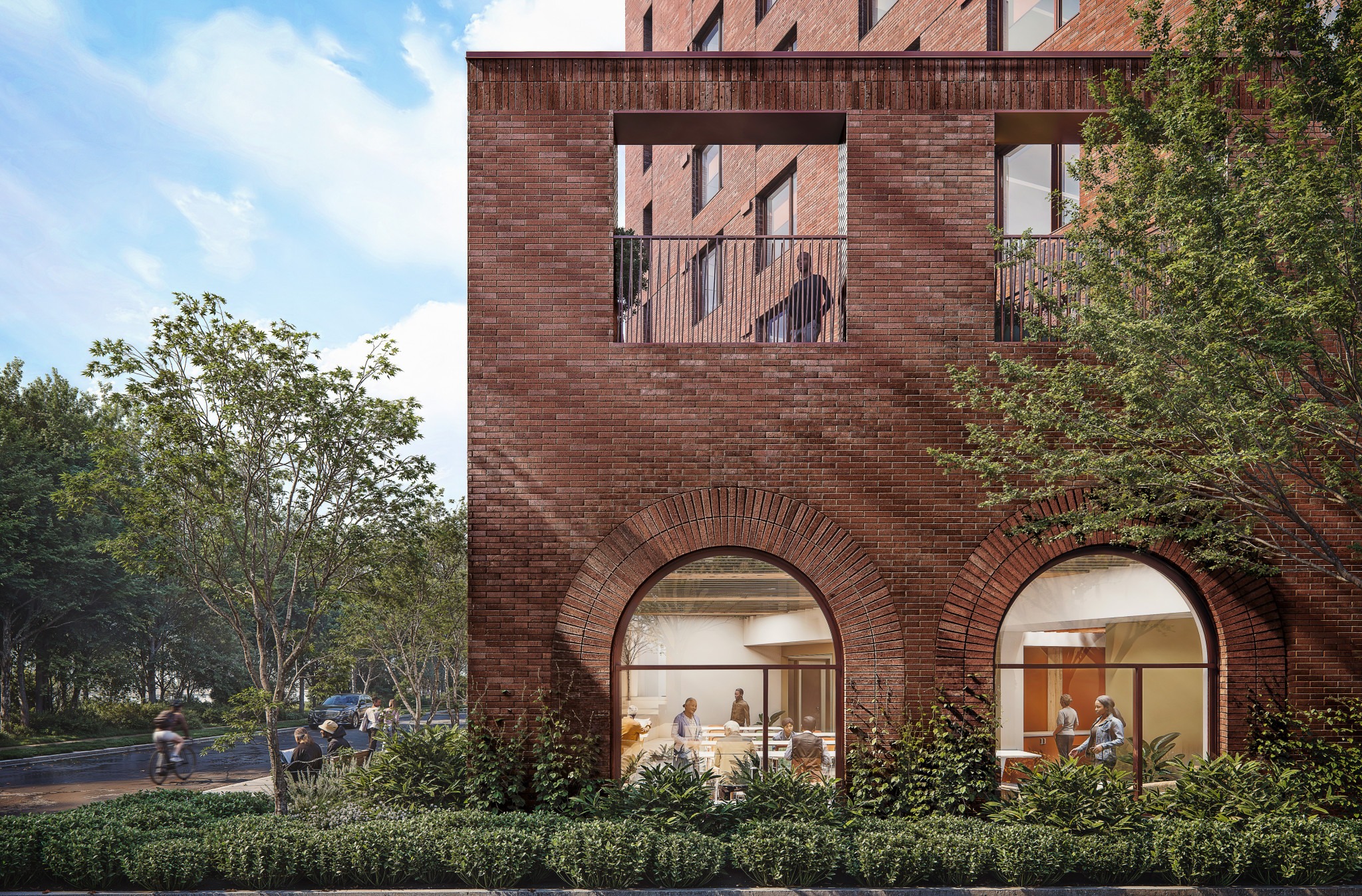 Red-brick building with arched windows, people visible inside, and landscaped greenery along a tree-lined street.