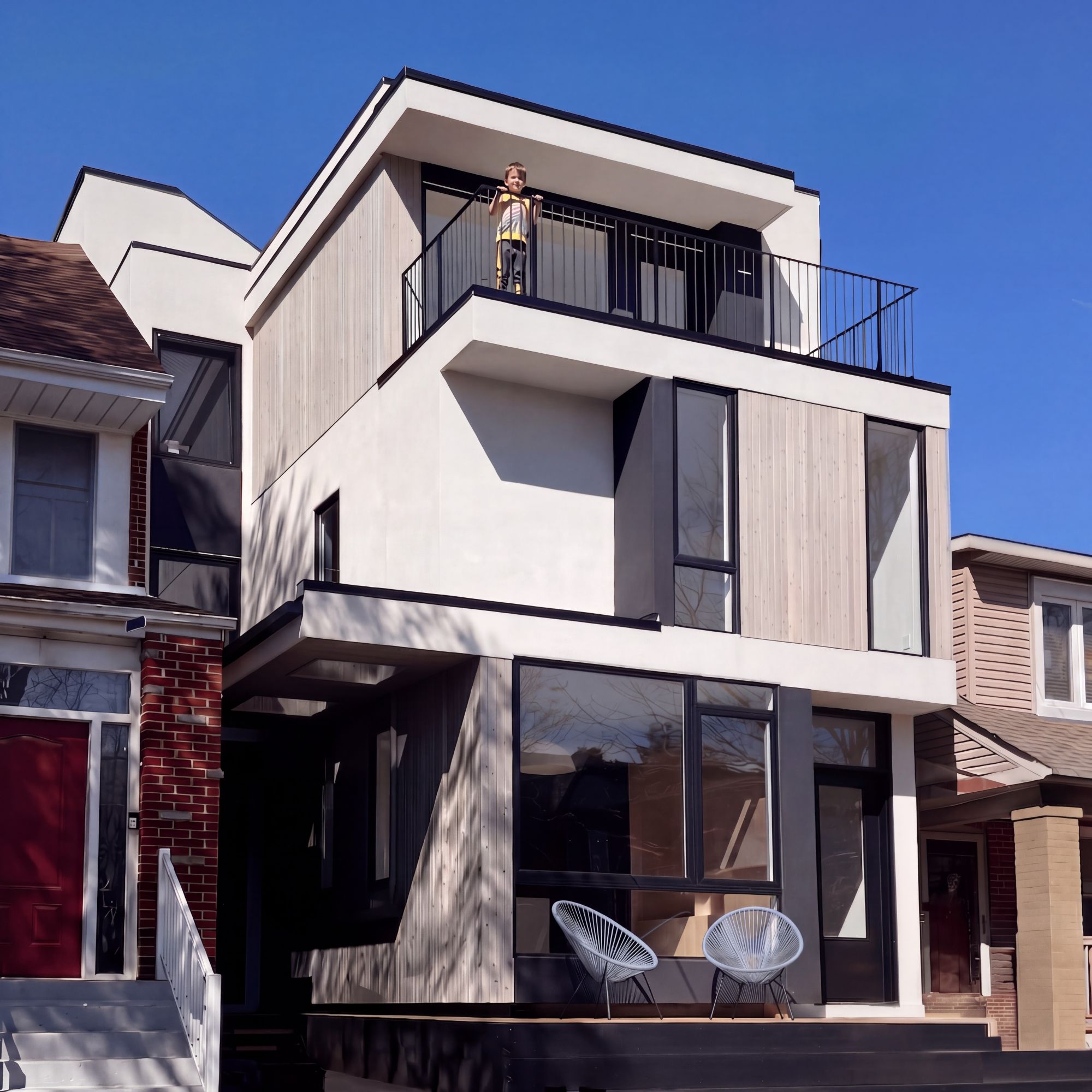 Modern townhouse with a child standing on the upper balcony and two white chairs on the lower deck.