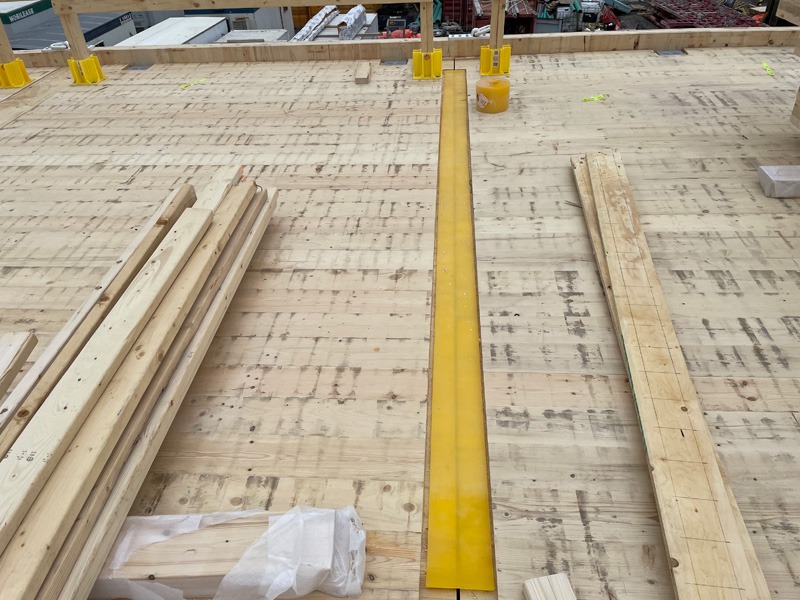 Construction site with a wooden deck, stacked lumber, yellow supports, and a long yellow beam running through the center.